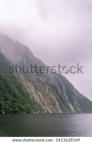 Dramatic waterfalls and cliffs at Milford Sound. Tourist attraction. View from boat cruise along Fjord. Rain and cloudy weather. Green and grey. Moody. South Island, New Zealand.
