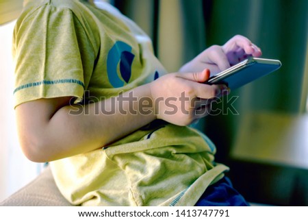 Cute little boy sitting in living room and playing mobile games. Close-up boy with smarthphone in his hands. Curious boy looking out the window. 