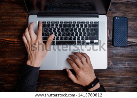 a male programmer in a black shirt is working at a laptop, close-up of the program code and developer's hands on a laptop. modern young programmer sitting at a wooden table at a computer