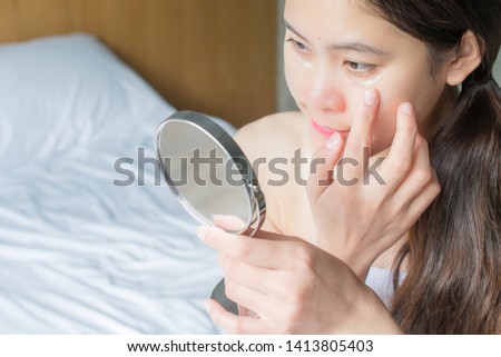 Portrait of Asian woman looking in the mirror and applying anti aging cream on her under eyes. The anti aging cream is using to reducing, masking or preventing signs of skin aging.