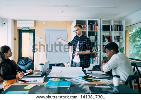 Young smart man in glasses standing at board with paper plan and talking to diverse colleagues standing in modern office room 