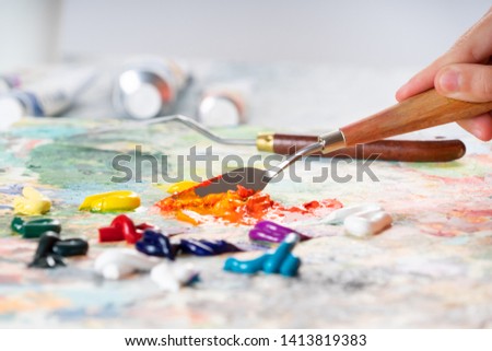 A young girl holds in his hand a palette-knife for drawing and mixes oiled paints. The process of mixing colors on the palette. Royalty-Free Stock Photo #1413819383