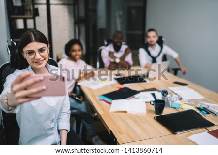 Happy caucasian female entrepreneur in optical spectacles for provide eyes protection spending time in office room taking selfie pictures via smartphone app with friendly colleagues on background