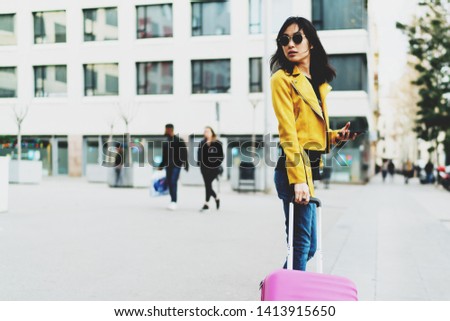 Side view photo of model look asian hipster female wearing sunglasses and modern outfit looking back while standing with a pink suitcase and a mobile phone in her hand on a blurred street background.