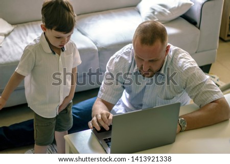 Happy young father sit on floor using laptop, relax with son and have fun together. Top view picture. Cute little boy standing next to his father, while using laptop. 