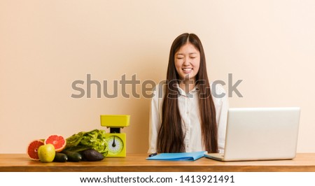 Young nutritionist chinese woman working with her laptop laughs and closes eyes, feels relaxed and happy.