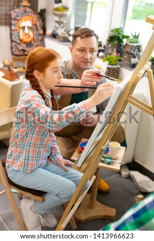 Coloring with teacher. Red-haired pupil sitting near drawing easel coloring picture with teacher