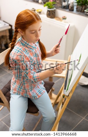 Coloring picture. Red-haired girl feeling joyful while coloring her picture holding painting brush