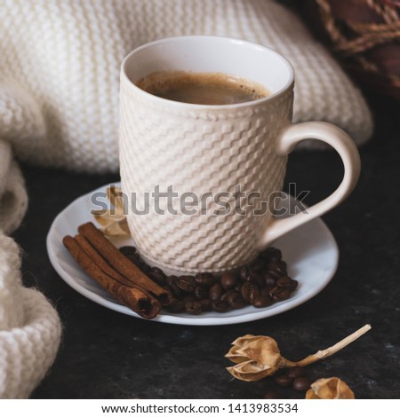 A cup of coffee is the key to a good mood. On a black, dark textural background, a composition of white, cream scarf and a white cup with espresso, coffee beans, cinnamon sticks and flowers. Square.
