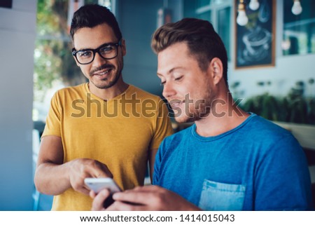 Half length portrait of cheerful male blogger in classic spectacles for provide eyes protection smiling at camera while friend standing near and reading positive comments near publication on website