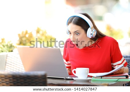 Amazed student e-learning with a laptop and headphones finds offer sitting in a coffee shop terrace