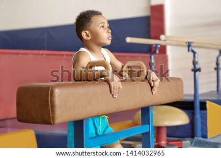 Picture of ten year old Afro American kid in white t-shirt standing by pommel horse at gym, having thoughtful facial expression, dreaming that he wins artistic gymnastics world champinonship