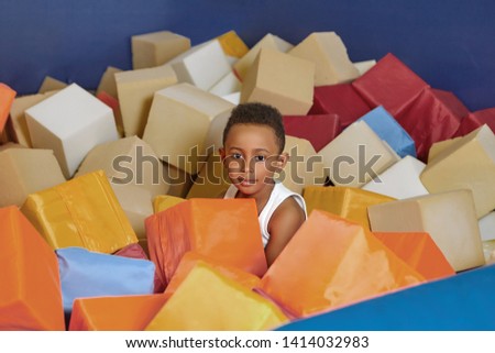 Happy African American eight year old boy playing with soft cubes in dry pool of children's room on birthday. Cute dark skinned child hiding among colorful cubes in trampoline of entertainment centre