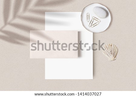 Tropical summer stationery mock-up scene. Blank business card, porcelain plate with stone, gold paper clips and sea shell, beige textured table background. Palm leaf shadow overlay. Flat lay, top view