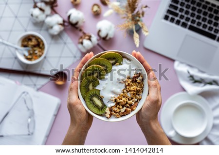 Female hand holding healthy breakfast bowl over work table background concept enjoy detox morning meal with laptop milk, woman eat natural granola nutrition detox food in home office, top view Royalty-Free Stock Photo #1414042814