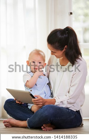 Young mother in casual clothes sitting on floor with baby boy on her laps and tablet computer in hands