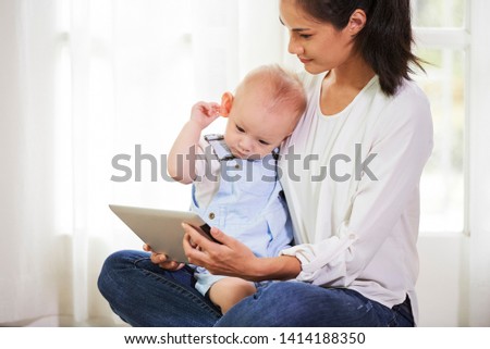Baby boy sitting on laptop of his mother, touching his ear and watching cartoon on tablet computer