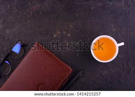 View from above. Fragrant tea in a white cup, beautiful flowers, a black background, a notebook with a pen and glasses. Office.