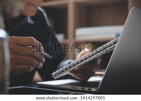 Asian business man holding pen reviewing business plan on notepad, working on laptop computer in office. Student reading content on notebook, studying online class, close up. E business, E learning