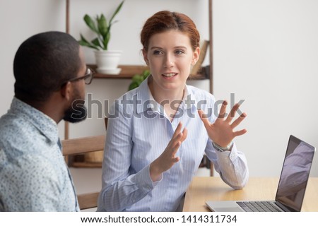 Two diverse businesspeople chatting sitting behind laptop in office. Excited caucasian female sharing ideas or startup business plan with black male coworker. Informal conversation, work break concept Royalty-Free Stock Photo #1414311734