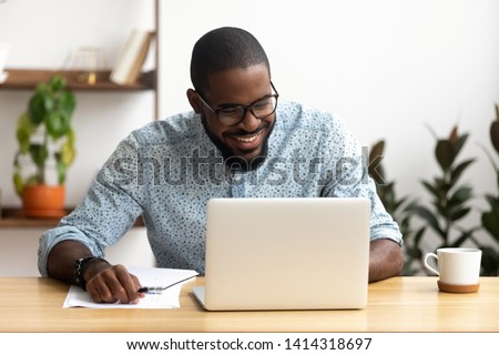 Smiling african-american manager sitting at office desk using laptop looking at screen. Handsome man reading good news, making funny video call, chatting social network. Coffee break concept Royalty-Free Stock Photo #1414318697