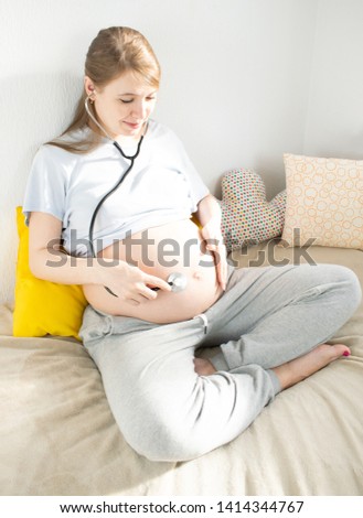 A young pregnant woman is holding a stethoscope near the belly. Listens to the heartbeat of the child. The concept of childbirth, prenatal medicine, newborn health.