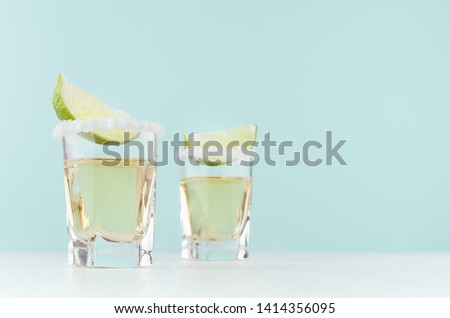 Tequila cocktail with salty rim, piece green lime in shot glasses on soft light pastel blue background, copy space.
