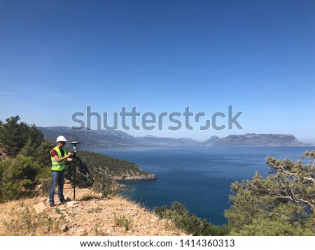 Geodetic engineer surveyor in white hard hat doing measurements with GNSS satellite receiver at the hill top above the sea Royalty-Free Stock Photo #1414360313