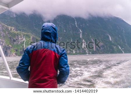 Man looking at beautiful waterfalls at Milford Sound. Tourist attraction. View from boat cruise along Fjord. Rain and cloudy weather. Green and grey. Moody. South Island, New Zealand