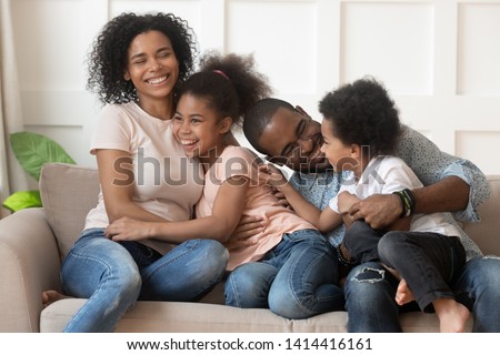Happy african american family of four laugh bond sit on sofa together, cheerful mixed race parents with kids embrace cuddle tickle on couch, young mom dad hug little cute children having fun at home