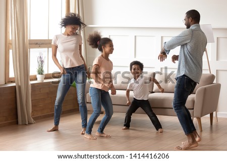 Happy funny active african american family of four parents and cute little mixed race kids dancing laughing in living room, black mom dad with small children having fun together enjoy leisure at home