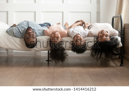 Happy african family bonding lying upside down on bed, young black parents and cute little kids posing for funny portrait in bedroom, mixed race mom dad with small children look at camera having fun