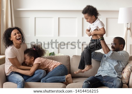 Happy black family having fun laughing with children, cheerful african american parents tickling lifting mixed race kids enjoy weekend on couch, mom dad with little son daughter playing at home