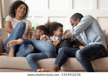 Happy cheerful african american family with cute little kids tickle laugh sit on sofa together, smiling young black parents and children having fun play at home enjoy funny lifestyle activity cuddle