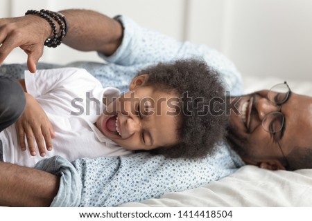 Cute adorable african american kid son having fun playing with father in bedroom, loving young black family dad tickling little child boy laughing bonding lying on bed together enjoy funny moment