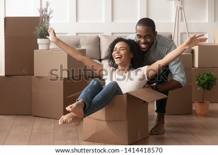 Happy african american young couple first time home buyers having fun unpacking laughing on moving day, excited wife riding sitting in cardboard box while black husband push it in new house apartment Royalty-Free Stock Photo #1414418570