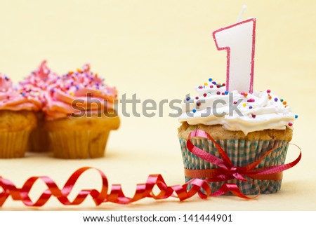Close-up shot of a cupcake tied with red ribbon and number 1 candle on top of it with blur view of strawberry cupcakes in background.