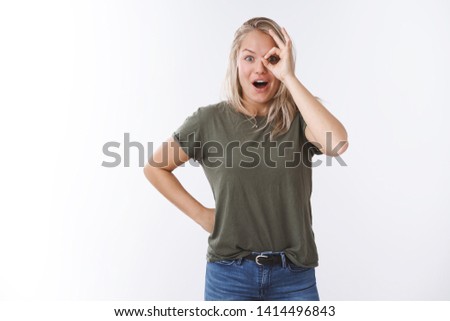 Surprised and amused beautiful caucasian blond in t-shirt showing okay sign over eye gasping dropping jaw astonished from amazing overwhelming promotion posing excited over white wall