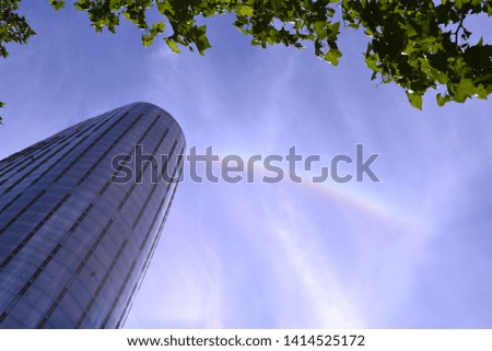 Amazing view of a skyscraper on blue sky, rainbow and green tree leaves background.