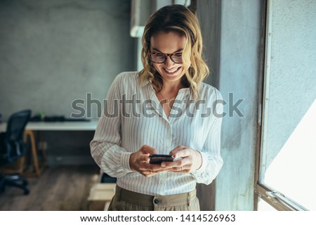 Businesswoman using her smart phone in office. Female entrepreneur looking at her mobile phone and smiling. Reading text messages. Royalty-Free Stock Photo #1414526963