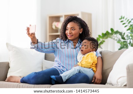 family, technology and motherhood concept - happy smiling young african american mother with little baby son taking selfie by smartphone at home