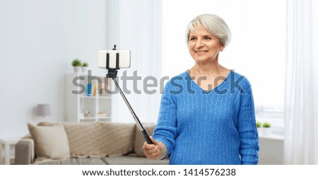 technology and old people concept - smiling senior woman taking picture by smartphone on selfie stick over home living room background