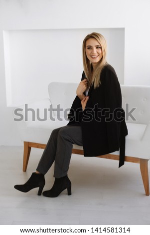Vertical photo of happy smiled blond young girl is wearing black suit, sitting on white sofa on white background. Place for text