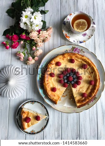 A cheesecake with blueberries and rasberries, a cup of tea with lemon and bunch of flowers on light wooden background. Beautiful delicious morning concept. Food blog recipe picture, sweets shop ad.
