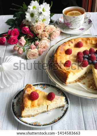 A cheesecake with blueberries and rasberries, a cup of tea with lemon and bunch of flowers on light wooden background. Beautiful delicious morning concept. Food blog recipe picture, sweets shop ad.