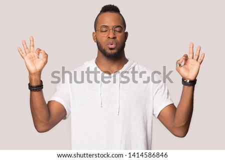 Stressed black guy meditating pose isolated on beige studio background, mixed race man in glasses folded fingers mudra symbol exhales and calms down breathing deep, self control, stress relief concept