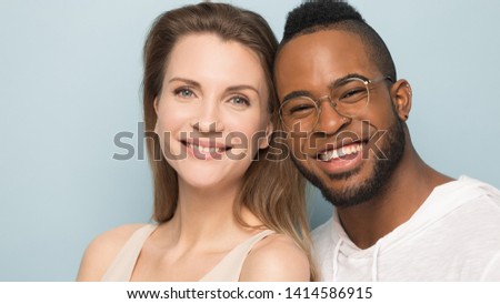 Headshot of smiling young caucasian woman next to beloved african american handsome millennial happy man in eyeglasses, diverse embracing couple posing for family photo on blue studio background.