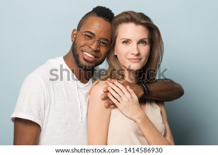 Happy handsome millennial african american guy in eyeglasses embracing smiling young pleasant caucasian woman. Attractive diverse couple posing together for family photo on blue studio background.