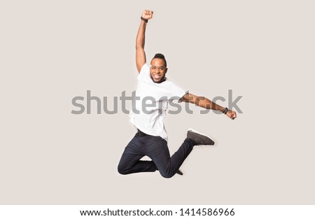 Happy joyful african american guy jumping high with raised hand and bent in knees legs between softbox lights on grey studio background, professional male model having fun during photo shooting