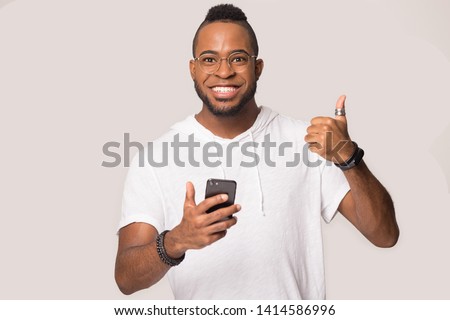 African man isolated on grey wearing white hoodie and glasses look at camera showing thumbs up holds smart phone feels happy, client satisfied by online sale, easy usage of e-commerce, new application